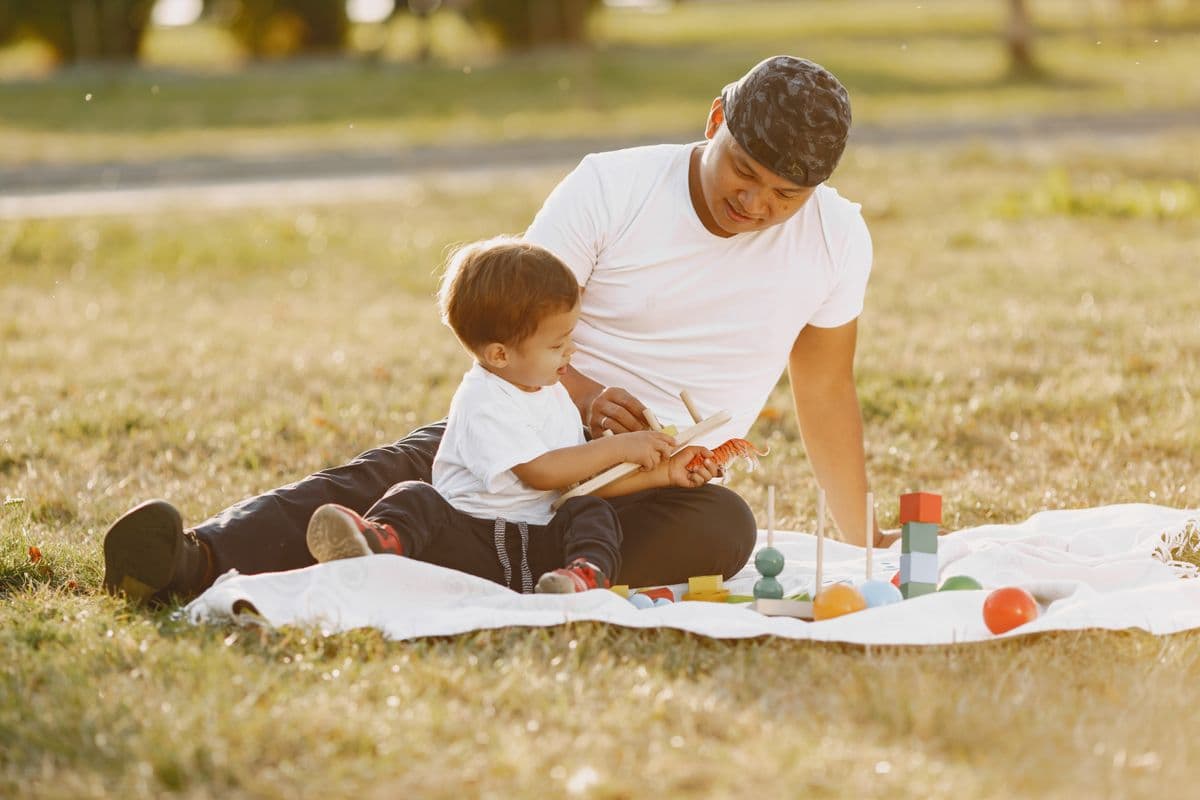 A father and his young son playing together on a blanket in a sunny park